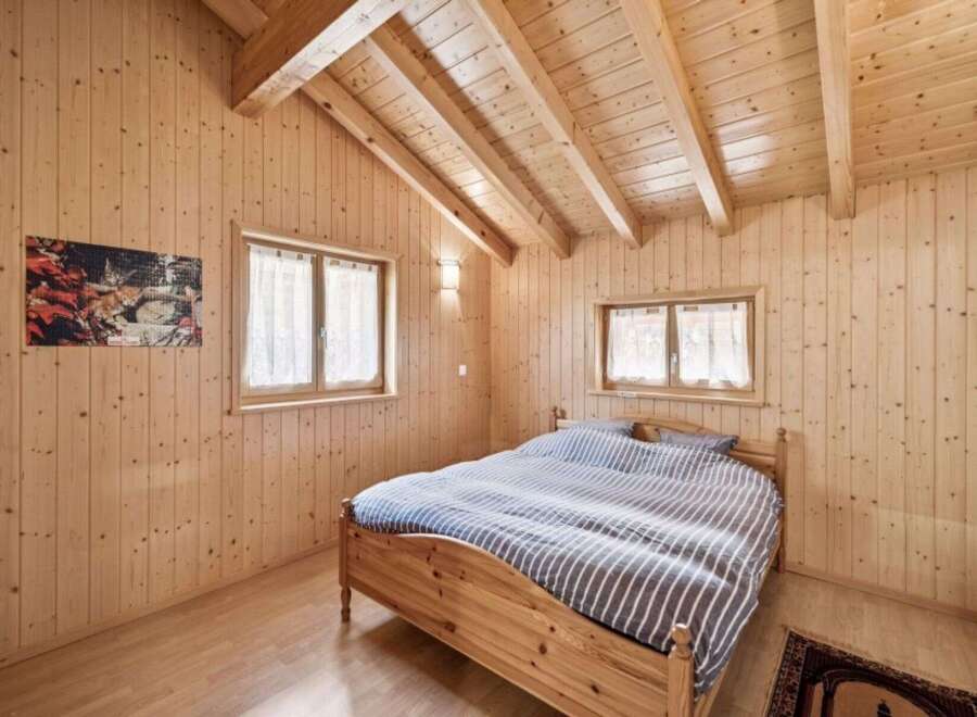 Experience cozy living in this charming wooden cabin bedroom, featuring a comfortable bed with striped bedding and natural light streaming through two windows.