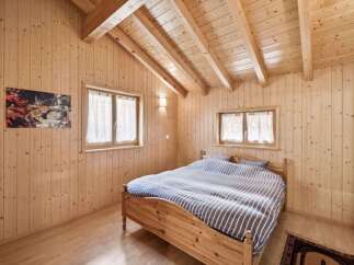Experience cozy living in this charming wooden cabin bedroom, featuring a comfortable bed with striped bedding and natural light streaming through two windows.