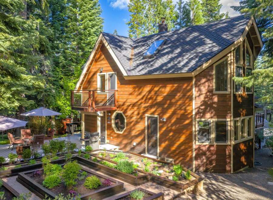 Discover this charming wooden home nestled amongst tall trees, featuring a multi-level deck with outdoor seating and umbrellas, perfect for enjoying the serene surroundings.