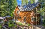 Discover this charming wooden home nestled amongst tall trees, featuring a multi-level deck with outdoor seating and umbrellas, perfect for enjoying the serene surroundings.