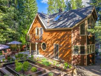 Discover this charming wooden home nestled amongst tall trees, featuring a multi-level deck with outdoor seating and umbrellas, perfect for enjoying the serene surroundings.