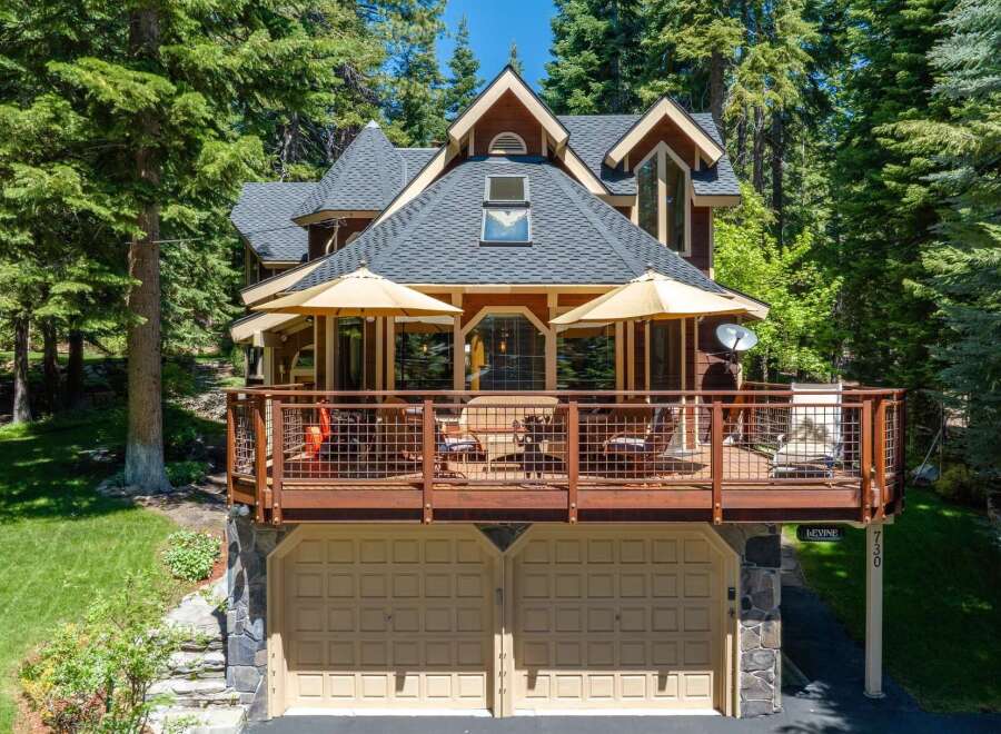 Nestled amidst tall pine trees, this charming home offers a spacious wooden deck perfect for outdoor entertaining, complete with umbrellas and seating.