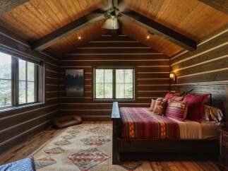 Experience rustic luxury in this charming log cabin bedroom, featuring a vaulted wood-paneled ceiling with exposed beams and a cozy king-sized bed adorned with a vibrant Southwestern-inspired comforter and pillows.