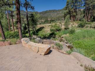 Enjoy the serene natural beauty of this property, featuring a unique stone bench on a paved patio overlooking a lush green landscape and a gentle stream.