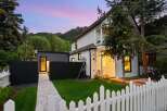 Charming white clapboard home with a white picket fence and lush green lawn, set against a backdrop of rolling hills and a vibrant twilight sky.