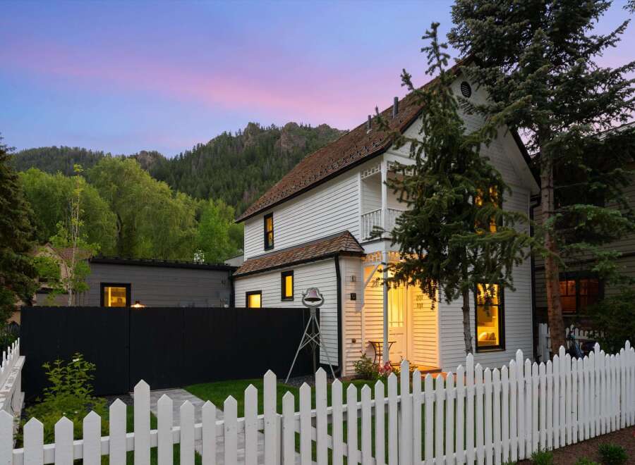 Charming white clapboard home with a white picket fence, nestled against a backdrop of lush green trees and majestic mountains.