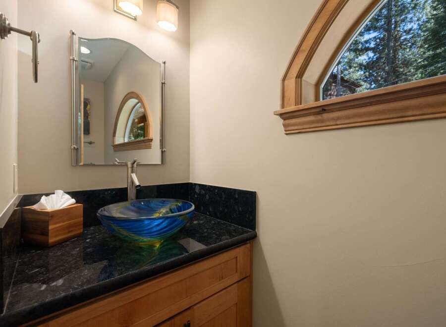 This powder room features a striking blue and green glass vessel sink atop a black granite countertop, complemented by warm wooden cabinetry.
