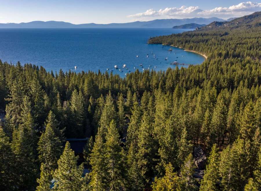 Experience the ultimate lakeside living with this stunning aerial view of a property nestled amidst a dense pine forest, offering breathtaking vistas of a crystal-clear blue lake.