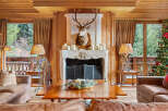 Experience rustic luxury in this charming living room, featuring a grand stone fireplace adorned with a majestic deer mount and surrounded by warm wood paneling.