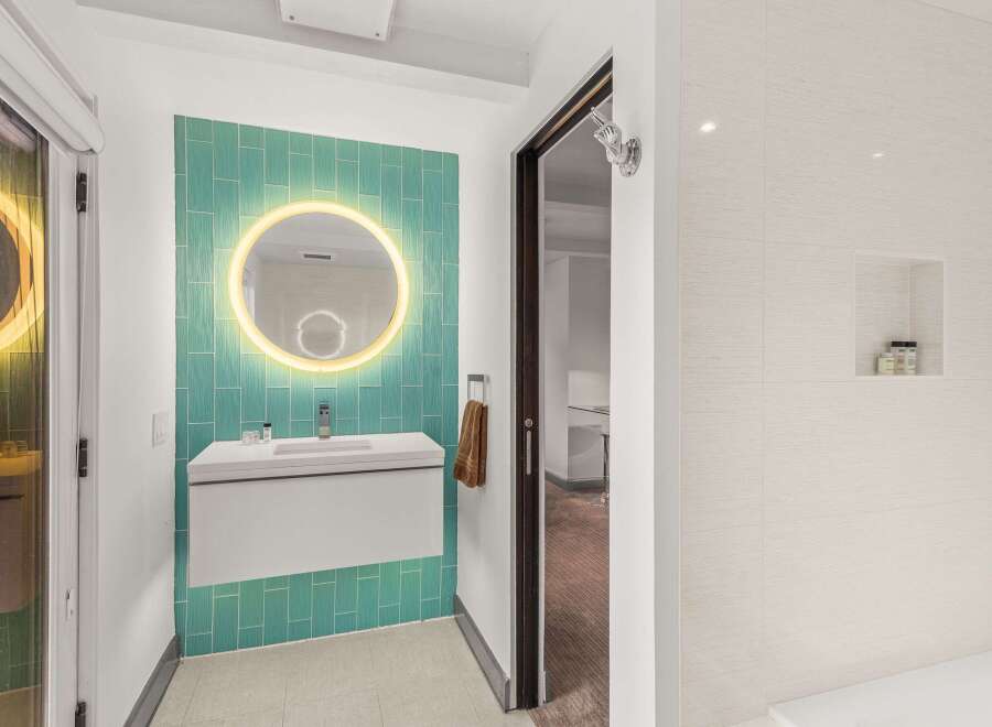 Experience modern luxury in this stunning bathroom, featuring a vibrant teal tiled accent wall with a backlit circular mirror.