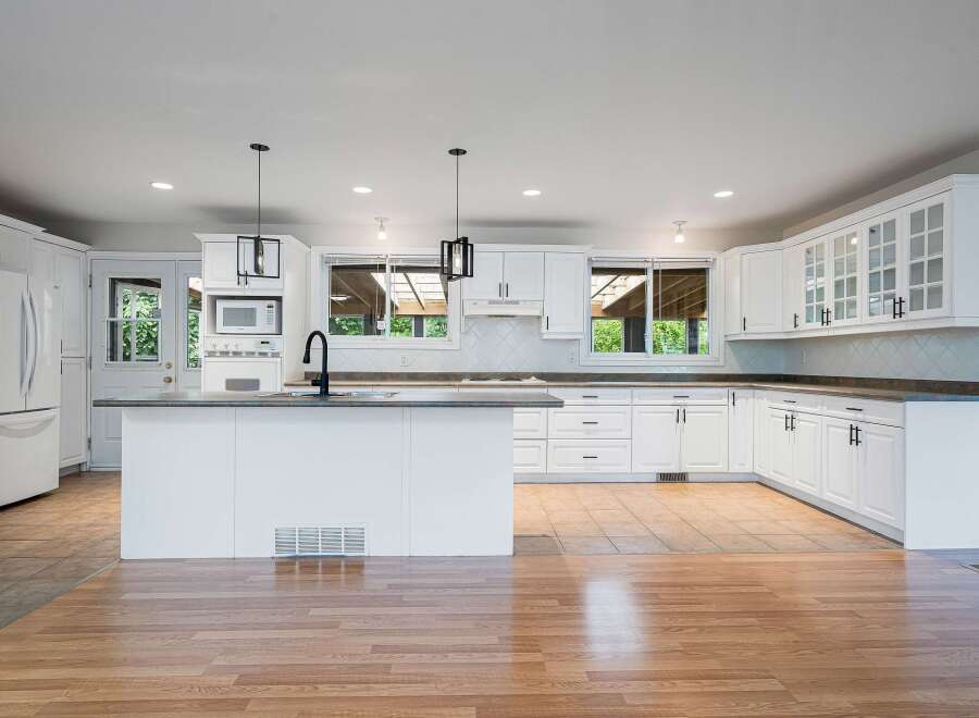 This spacious kitchen features pristine white cabinetry, a large central island with a sink and faucet, and a sleek tiled backsplash.