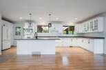 This spacious kitchen features pristine white cabinetry, a large central island with a sink and faucet, and a sleek tiled backsplash.