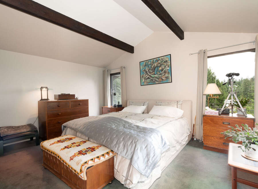 Experience serene living in this charming bedroom featuring vaulted ceilings with exposed dark wood beams and a large window offering picturesque forest views.
