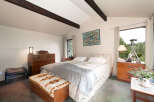 Experience serene living in this charming bedroom featuring vaulted ceilings with exposed dark wood beams and a large window offering picturesque forest views.