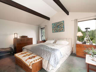 Experience serene living in this charming bedroom featuring vaulted ceilings with exposed dark wood beams and a large window offering picturesque forest views.