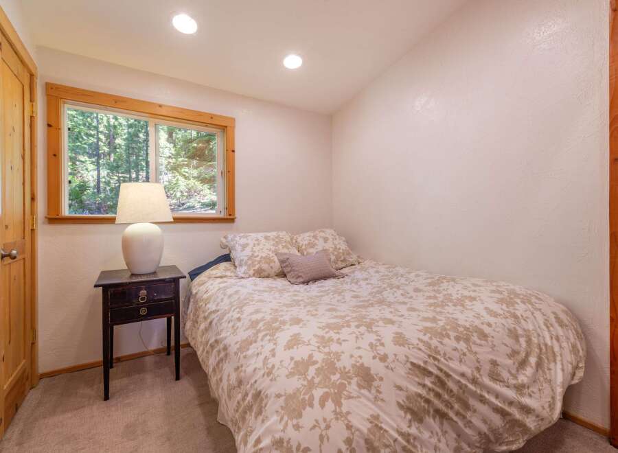 This cozy bedroom features a comfortable bed with a floral comforter, a bedside table with a lamp, and a window offering serene forest views.