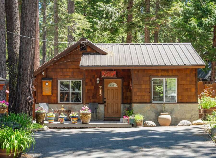 Charming wooden cabin nestled amongst tall trees, featuring a welcoming porch adorned with potted plants and a rustic stone foundation.