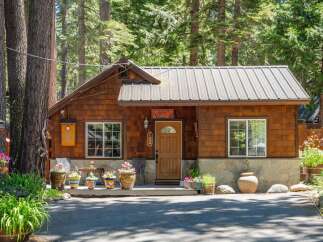 Charming wooden cabin nestled amongst tall trees, featuring a welcoming porch adorned with potted plants and a rustic stone foundation.