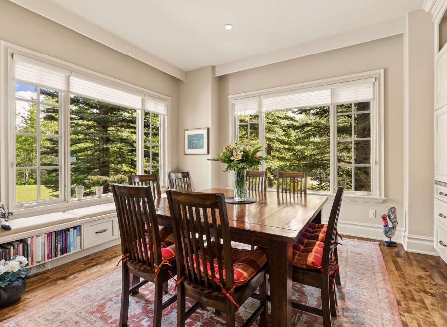 Enjoy meals with a view in this charming dining room, featuring a large wooden table and chairs set against a backdrop of lush greenery through expansive windows.
