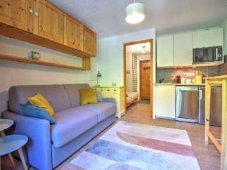 This cozy studio apartment features a comfortable sofa, a compact kitchen area with modern appliances, and ample storage with overhead wooden cabinets.