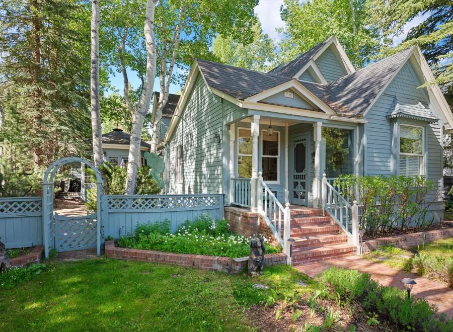 Charming light blue home with a welcoming front porch and white railings, nestled amongst mature trees.