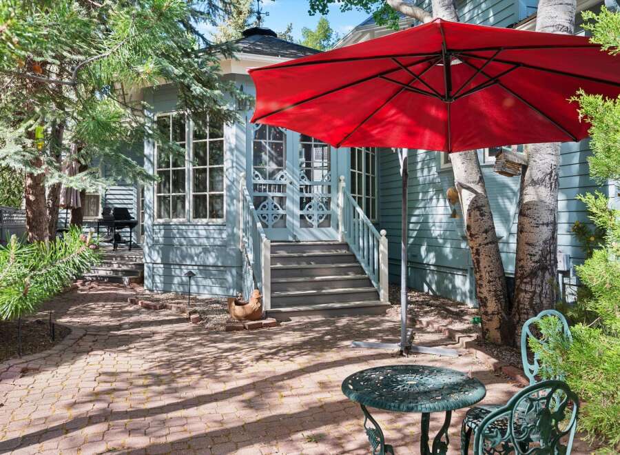 Enjoy outdoor living in this charming backyard featuring a red umbrella, a wrought iron table and chairs, and a brick patio perfect for al fresco dining.