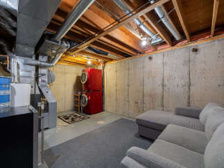 Discover the potential of this unfinished basement, featuring a stacked red washer and dryer set and a comfortable grey sectional sofa.