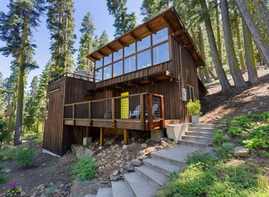 Nestled amidst tall trees, this charming wooden cabin offers a unique architectural design with a striking wall of windows on the upper level, flooding the interior with natural light.