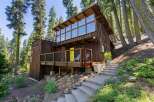 Nestled amidst tall trees, this charming wooden cabin offers a unique architectural design with a striking wall of windows on the upper level, flooding the interior with natural light.