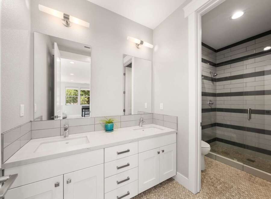 This modern bathroom features a spacious double vanity with white quartz countertops and sleek cabinetry, complemented by a stylish tiled shower enclosure with horizontal gray and white accents.
