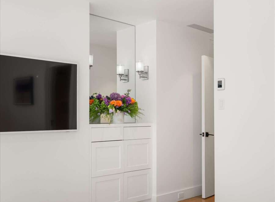 Experience modern luxury in this beautifully appointed bedroom, featuring a sleek wall-mounted television and elegant sconce lighting above a charming floral arrangement.
