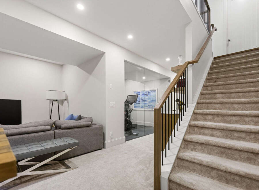 Discover modern living with this spacious basement featuring a comfortable grey sectional sofa and a dedicated home gym enclosed by sleek glass walls.