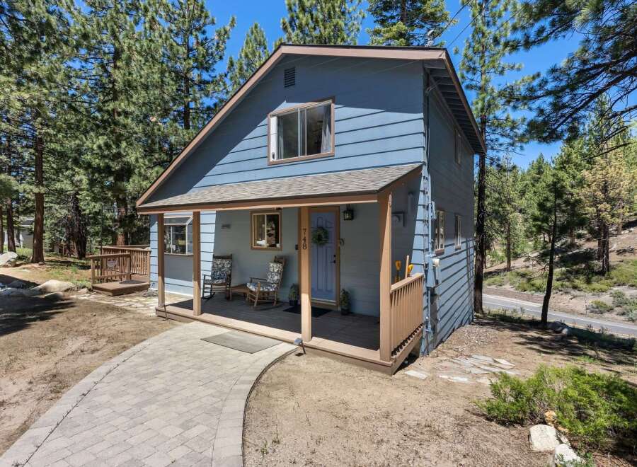 Charming blue home nestled among tall pine trees, featuring a welcoming covered porch with rocking chairs and a paved walkway leading to the entrance.