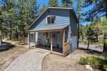 Charming blue home nestled among tall pine trees, featuring a welcoming covered porch with rocking chairs and a paved walkway leading to the entrance.