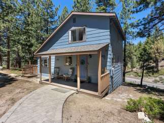 Charming blue home nestled among tall pine trees, featuring a welcoming covered porch with rocking chairs and a paved walkway leading to the entrance.
