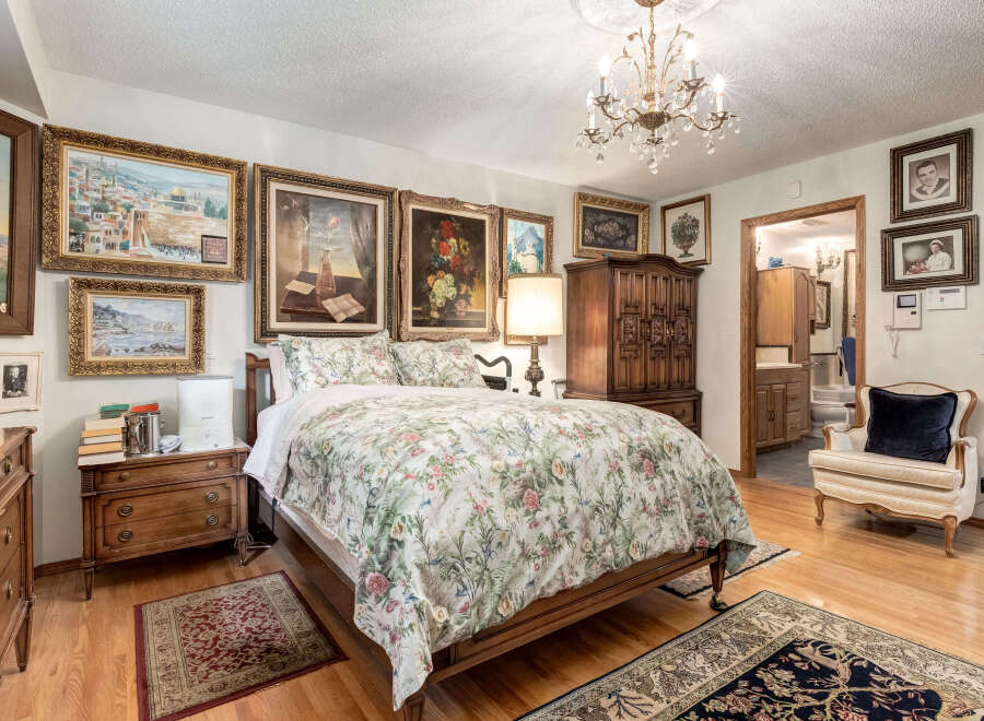 This beautifully decorated bedroom features a luxurious floral comforter on a wooden bed frame, complemented by an ornate chandelier and a collection of framed artwork adorning the walls.