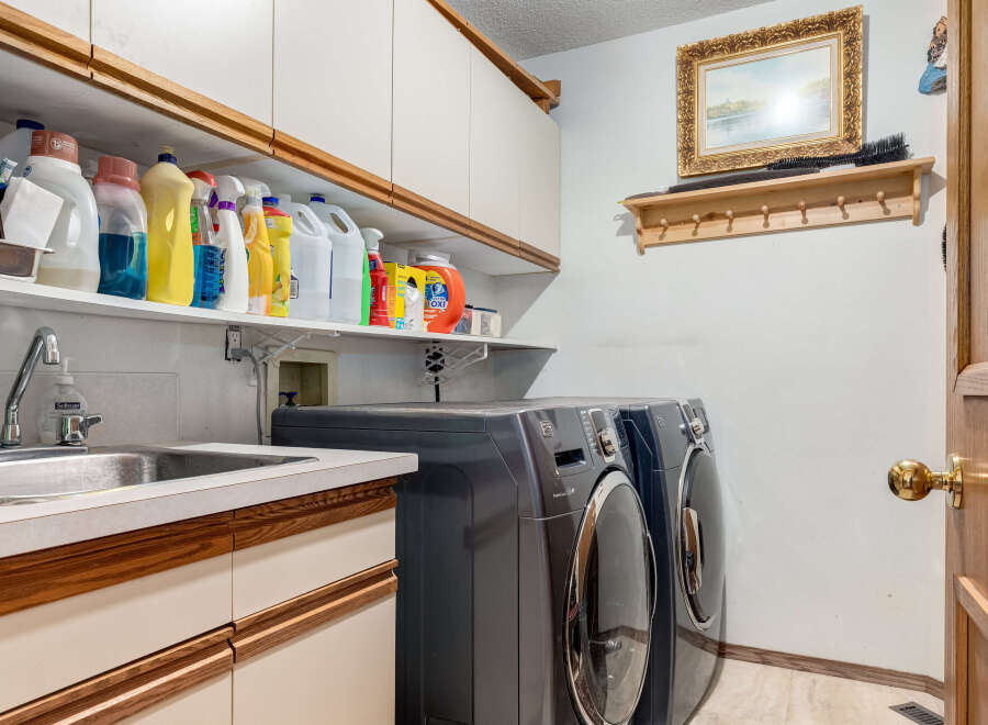 This laundry room features a convenient sink and ample storage with overhead cabinets and open shelving stocked with cleaning supplies.