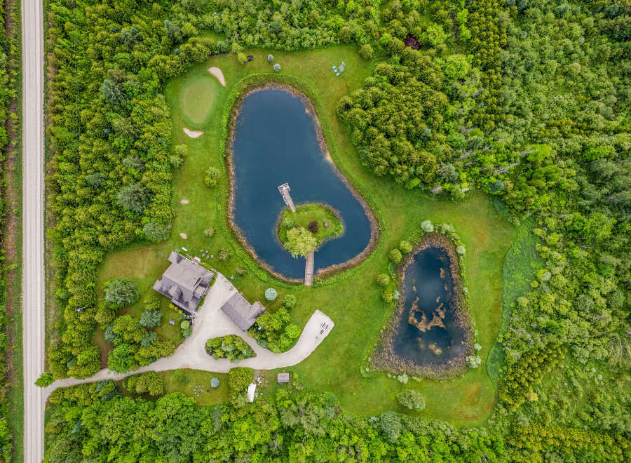 Discover this stunning aerial view of a private estate featuring two serene ponds, a charming golf putting green, and a picturesque island accessible by a wooden bridge.