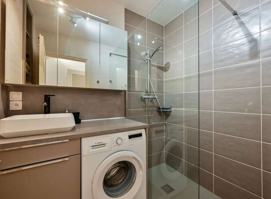 This modern bathroom features a sleek vessel sink atop a taupe vanity, complemented by a built-in Bosch washing machine.
