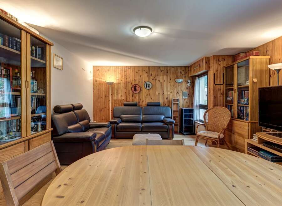 This inviting living room features a warm, wood-paneled ambiance, complemented by comfortable leather sofas and a large, light-wood dining table perfect for gatherings.