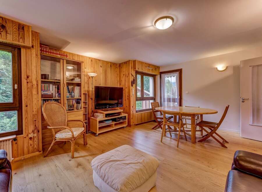 Experience cozy living in this charming wood-paneled living room, featuring a comfortable seating area with a plush ottoman and a dedicated dining space with a round wooden table and chairs.