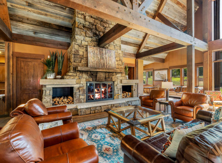 Experience rustic luxury in this stunning living room featuring a grand stone fireplace with a crackling fire, complemented by exposed wooden beams and a vaulted ceiling.