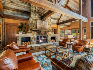 Experience rustic luxury in this stunning living room featuring a grand stone fireplace with a crackling fire, complemented by exposed wooden beams and a vaulted ceiling.