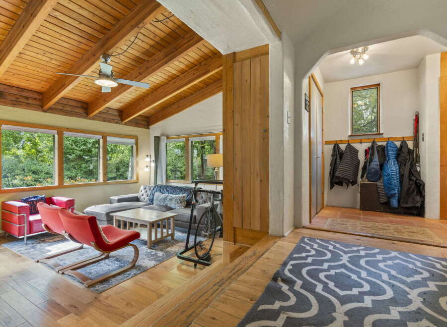 Step into a charming living space featuring a vaulted wood-beamed ceiling with a ceiling fan, complemented by large windows offering lush green views.