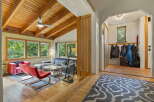 Step into a charming living space featuring a vaulted wood-beamed ceiling with a ceiling fan, complemented by large windows offering lush green views.