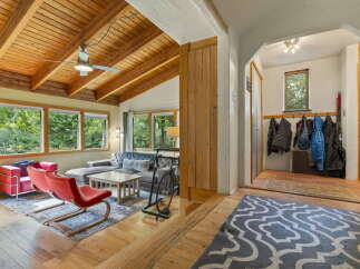 Step into a charming living space featuring a vaulted wood-beamed ceiling with a ceiling fan, complemented by large windows offering lush green views.