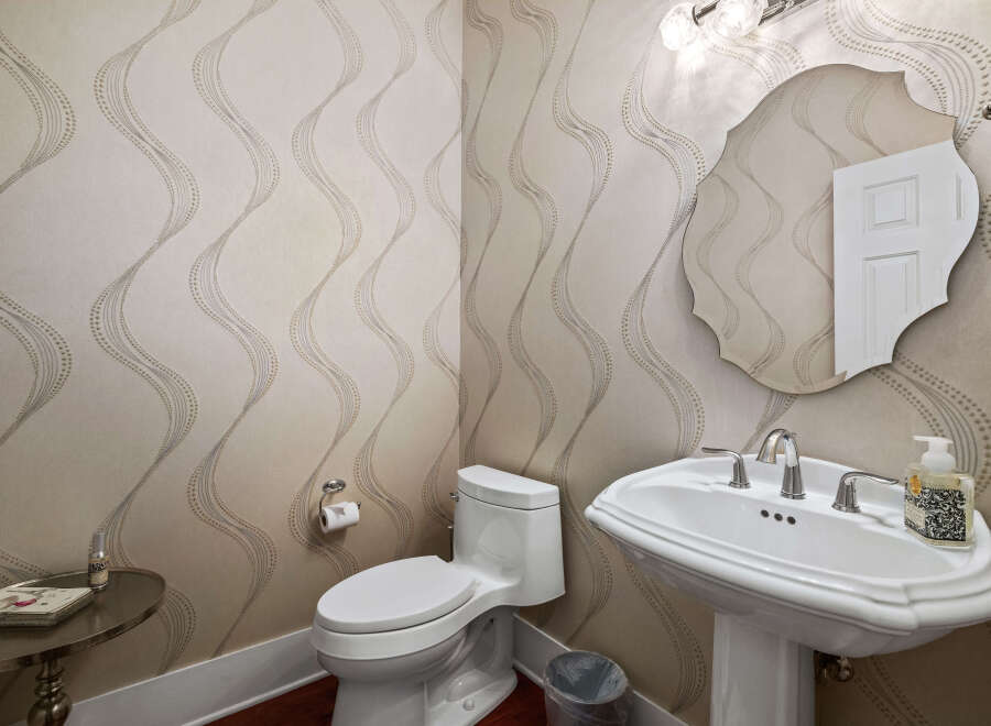 This powder room features elegant wallpaper with a subtle wave pattern, complemented by a classic white pedestal sink and a decorative mirror.