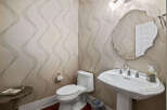 This powder room features elegant wallpaper with a subtle wave pattern, complemented by a classic white pedestal sink and a decorative mirror.