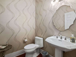 This powder room features elegant wallpaper with a subtle wave pattern, complemented by a classic white pedestal sink and a decorative mirror.