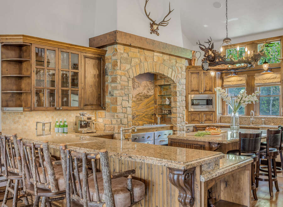Experience rustic elegance in this stunning kitchen, featuring a grand stone fireplace with a mounted deer head and a unique antler chandelier.